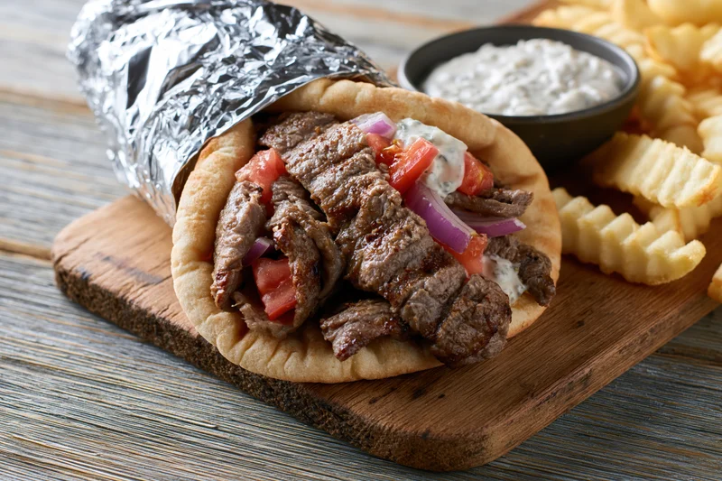 Gyro wrap with grilled beef, tomatoes, onions and tzatziki sauce on wooden board with crinkle-cut fries and dipping bowl