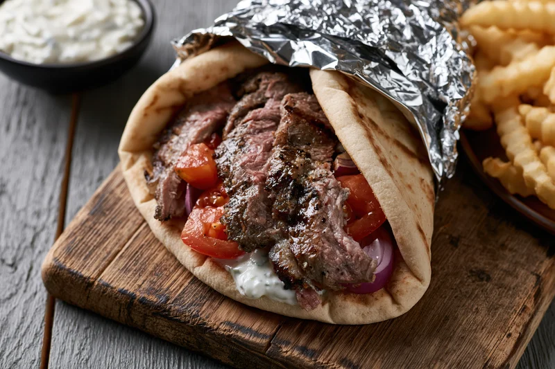 Grilled beef gyro in pita bread with tomatoes and onions, partially wrapped in foil, served on wooden board with tzatziki and fries