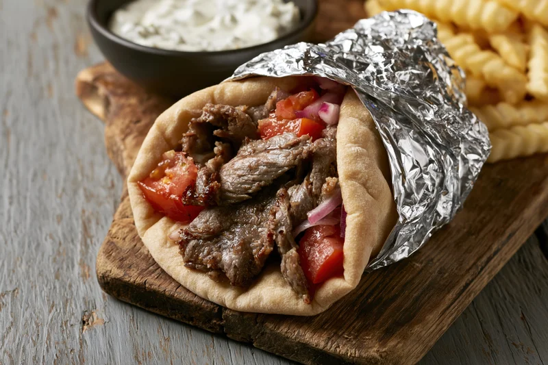 Gyro wrap with grilled meat, tomato, and onion on wooden board, partially wrapped in aluminum foil