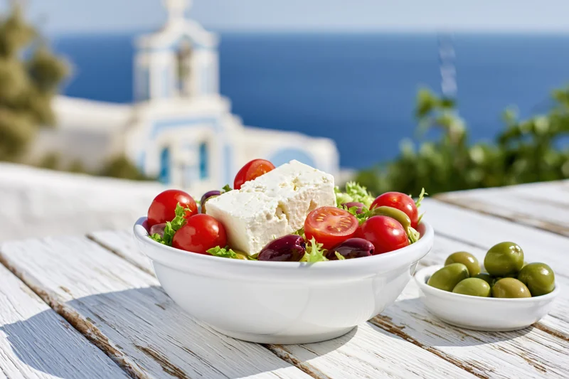 Greek salad with feta cheese, tomatoes, and olives in a white ceramic bowl on a wooden table overlooking a coastal church and sea