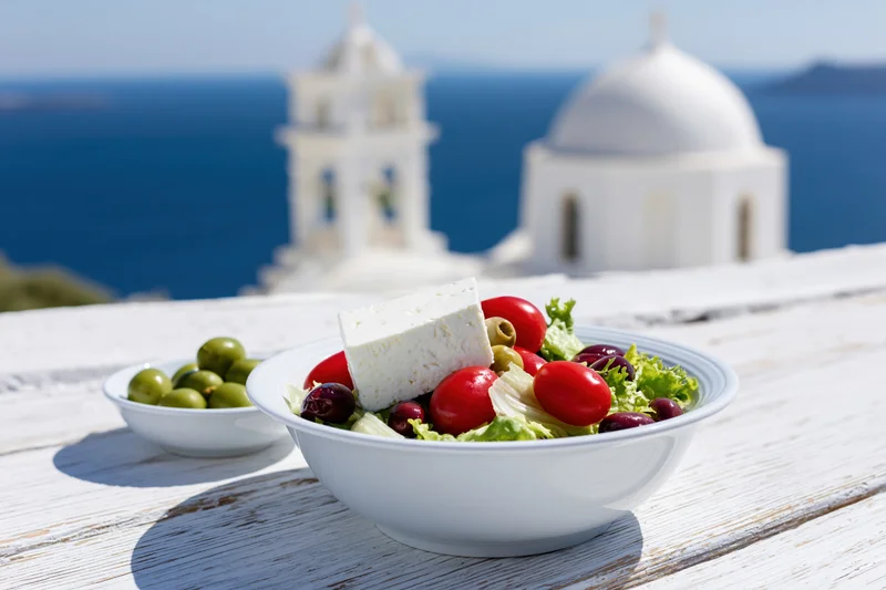 Greek salad with feta cheese, tomatoes, olives and lettuce in a white ceramic bowl on a wooden table overlooking a whitewashed church and the sea