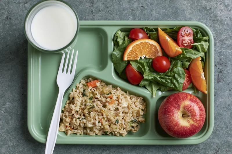 Green plastic school lunch tray with compartments of rice, salad, apple, orange, and milk, with white fork on gray surface