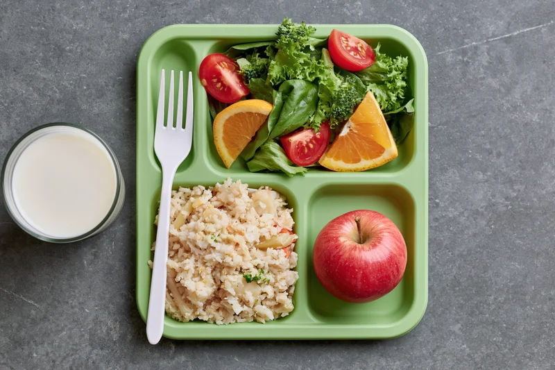Green plastic school lunch tray with compartments of rice, salad, apple and milk on gray surface
