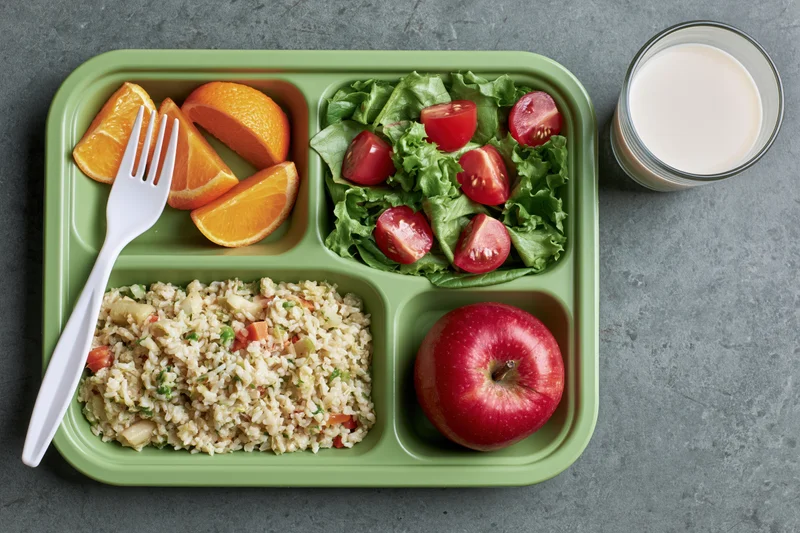 Green plastic school lunch tray with compartments of rice, salad, fruit and a glass of milk on a gray surface