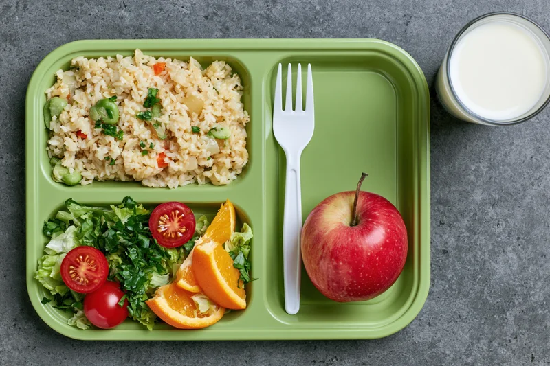 Green plastic school lunch tray with compartments of rice, vegetables, fruit and milk on gray surface