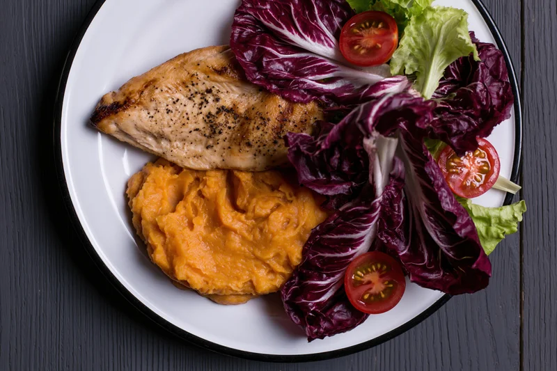 Grilled chicken breast with mashed sweet potato and mixed greens on a white plate with black rim on dark wood surface