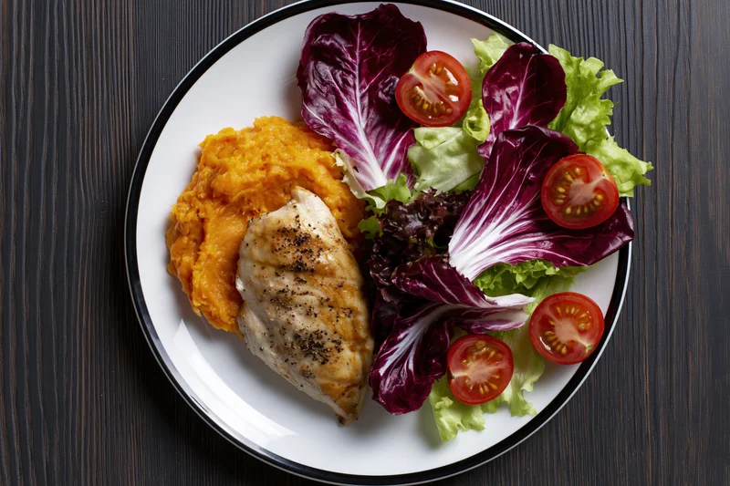 Grilled chicken breast with mashed sweet potato and mixed greens on a white plate with black rim, top view on dark wooden table