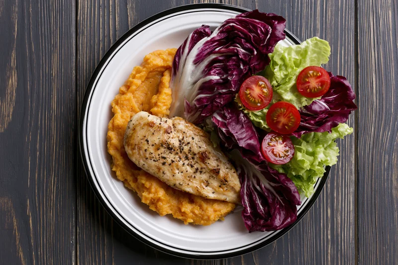 Grilled chicken breast with mashed sweet potato and mixed greens on a white plate with black rim on dark wooden table