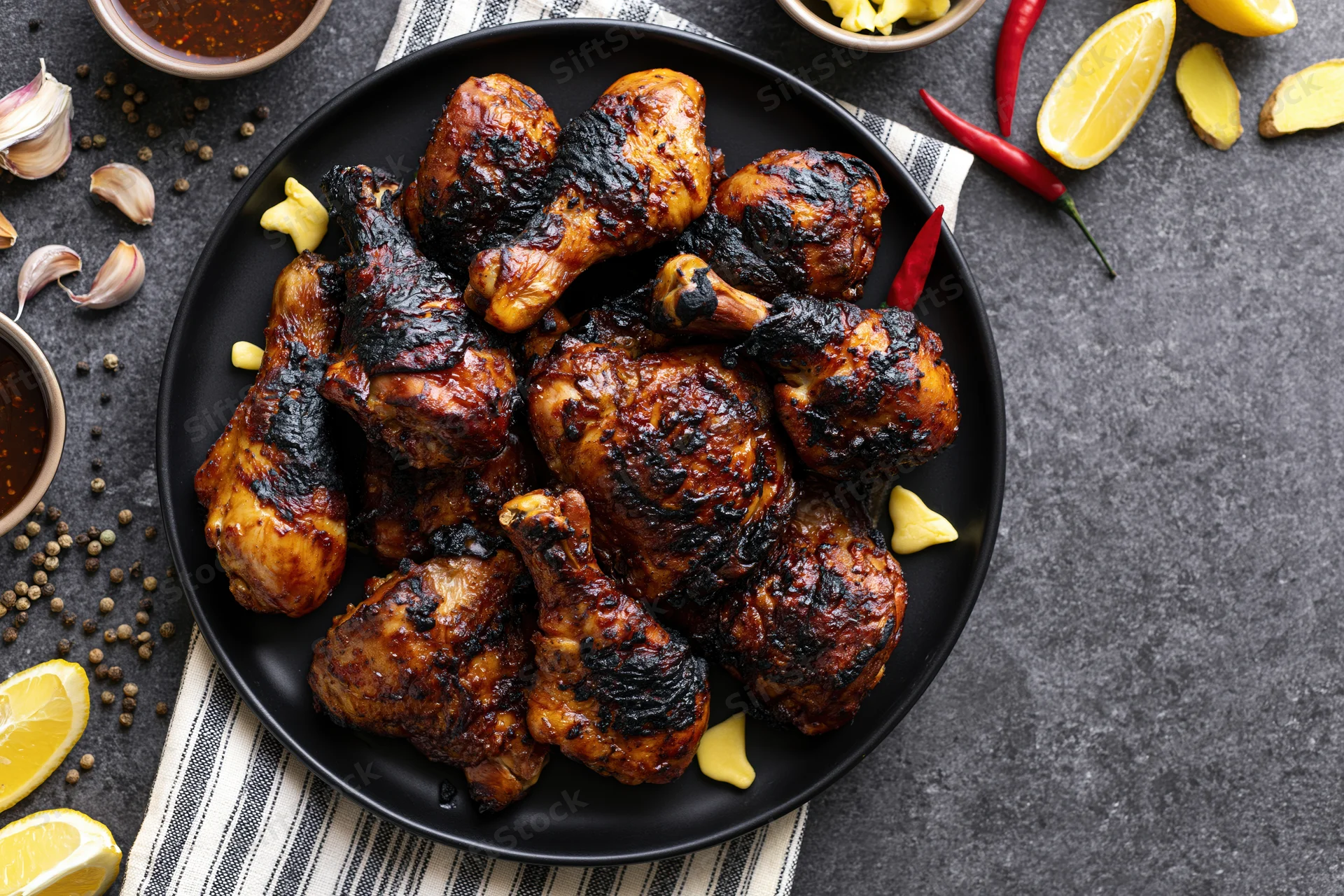 Grilled chicken legs and thighs piled on a black plate with lemon, chili, garlic, and spices around on dark surface