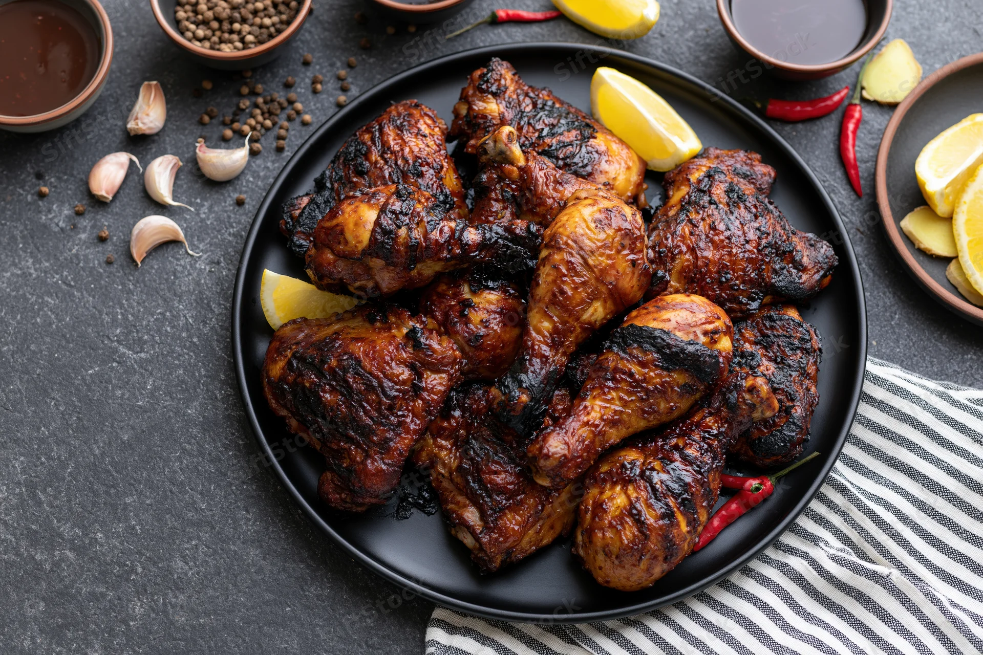 Grilled chicken legs and thighs piled on a black plate with lemon, chili, garlic, and spices around on dark surface