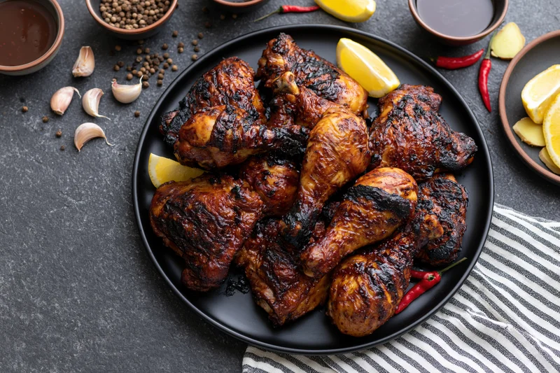 Grilled chicken legs and thighs piled on a black plate with lemon, chili, garlic, and spices around on dark surface
