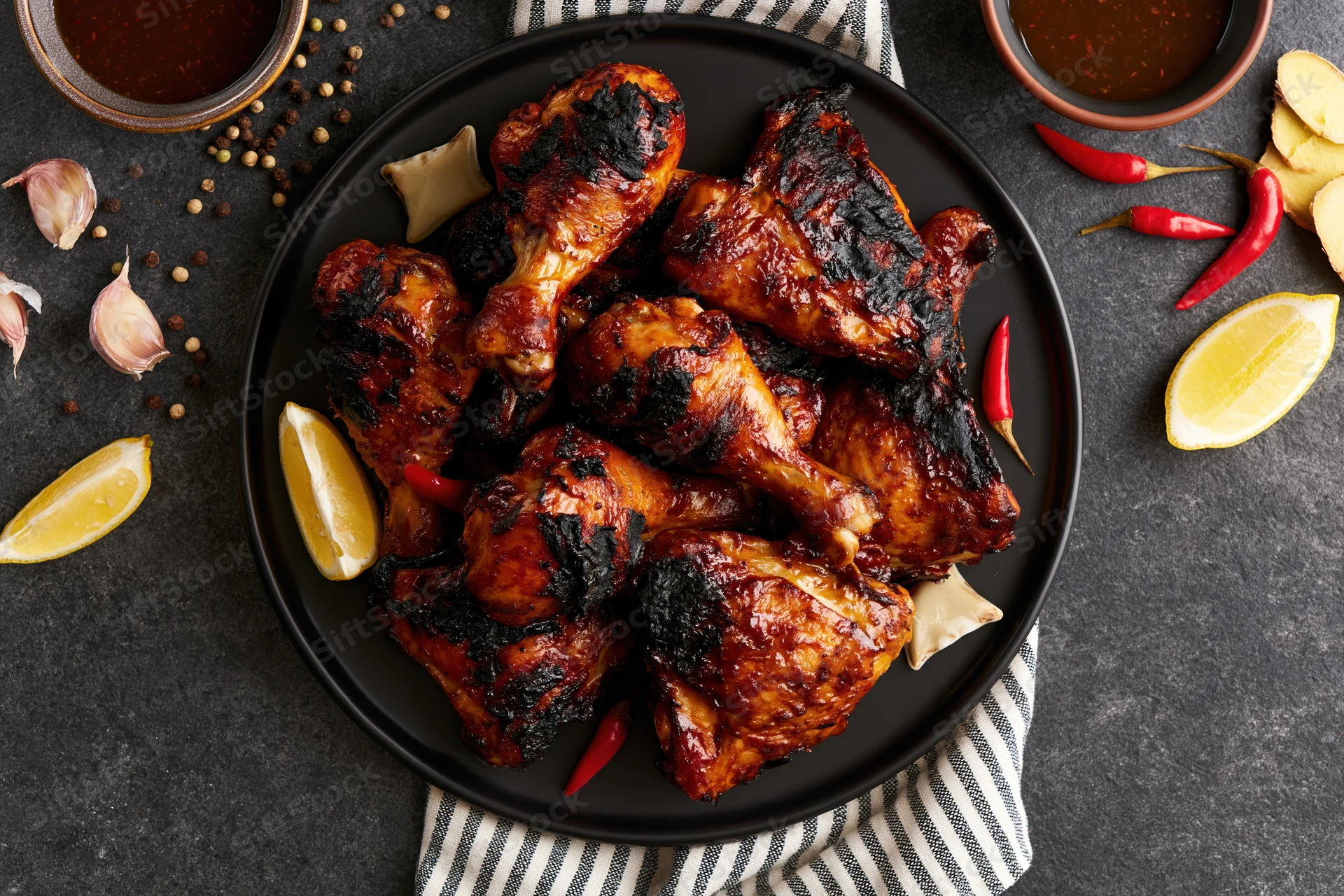 Grilled chicken legs and thighs piled on a black plate with lemon, chili, garlic, and spices around