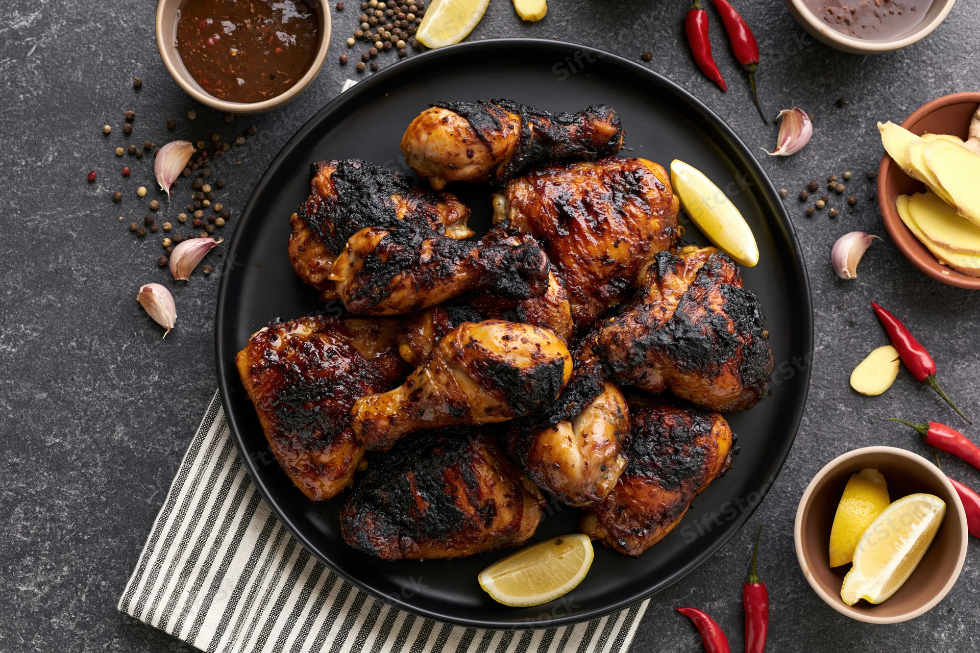 Grilled chicken legs and thighs piled on a black plate with lemon, chili, garlic, and spices around