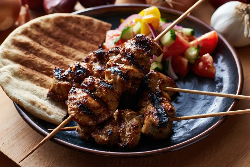 Grilled chicken skewers on wooden sticks served with flatbread and vegetable salad on a dark plate