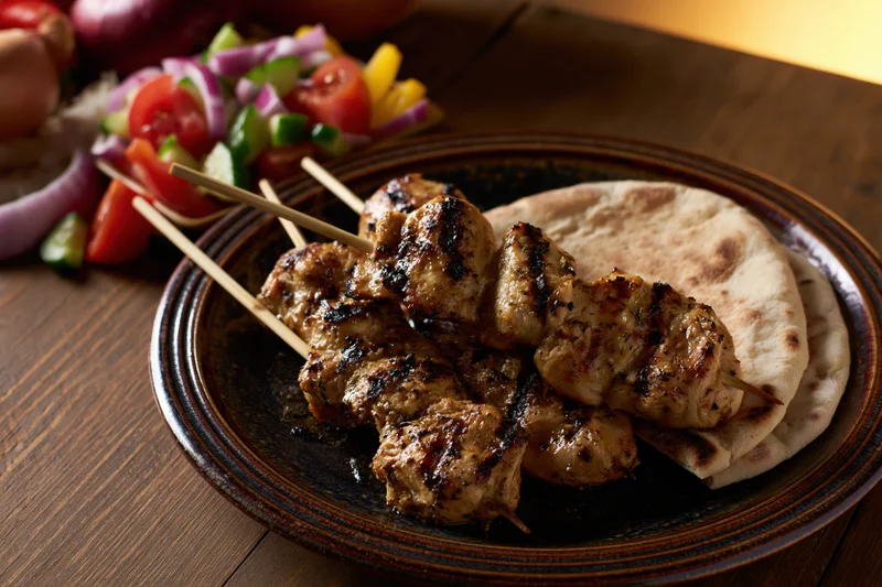 Grilled chicken skewers on a dark ceramic plate with flatbread and vegetable salad on a wooden table