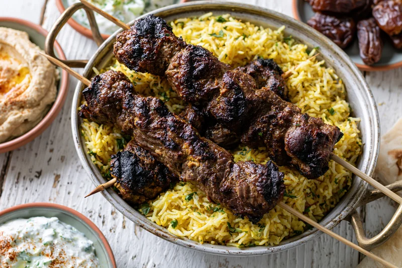 Grilled lamb kebabs on saffron rice in a ceramic bowl with side dishes on a rustic wooden table