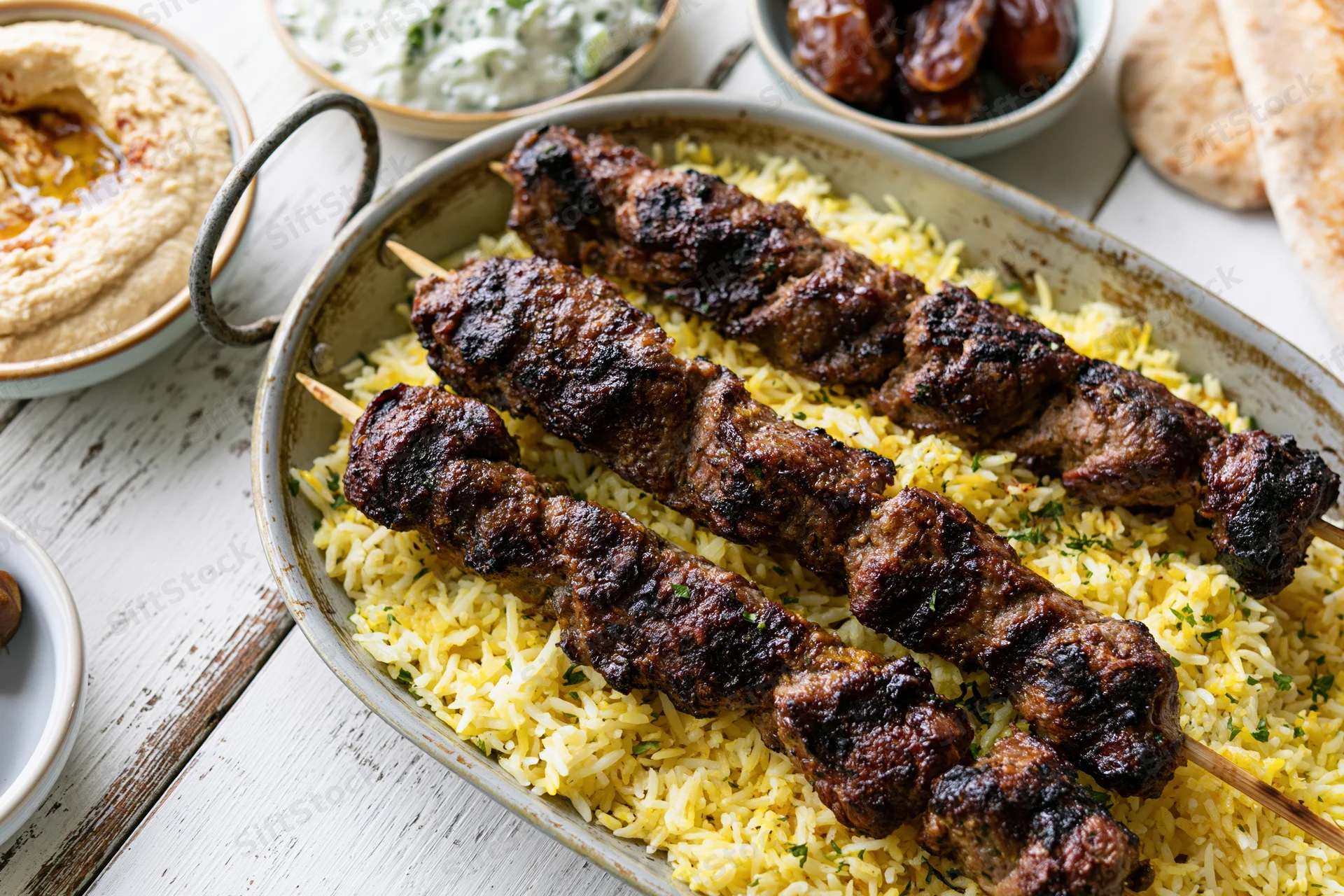 Grilled lamb kebabs on saffron rice in a metal tray with hummus, tzatziki, dates, and pita bread on a white wooden table