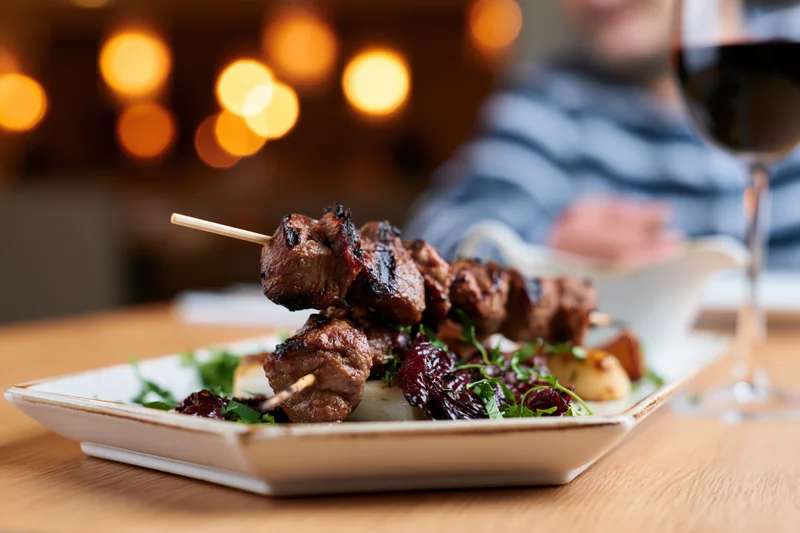 Grilled meat skewers on a white ceramic plate with garnish and wine glass on wooden table