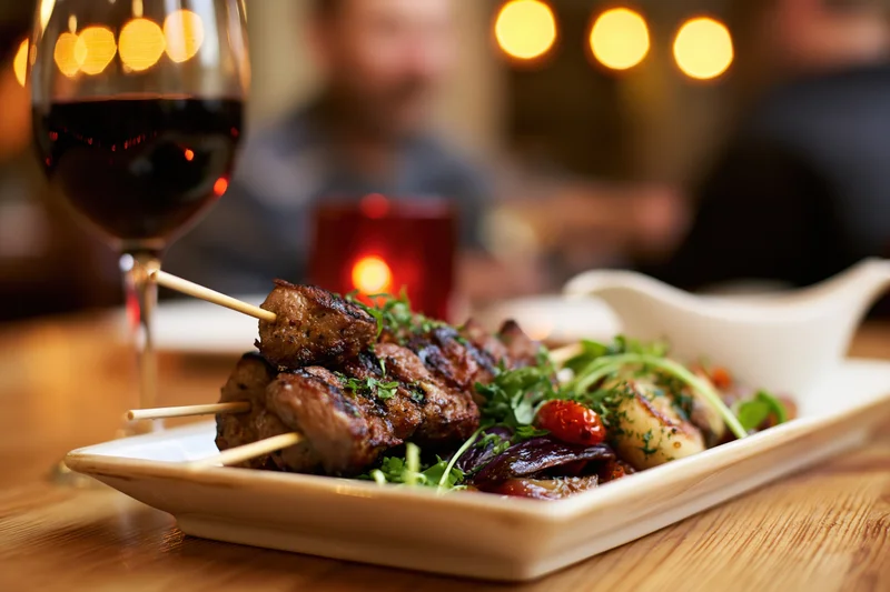 Grilled meat skewers on a white ceramic plate with herbs and vegetables, served with red wine and candlelight at a restaurant table