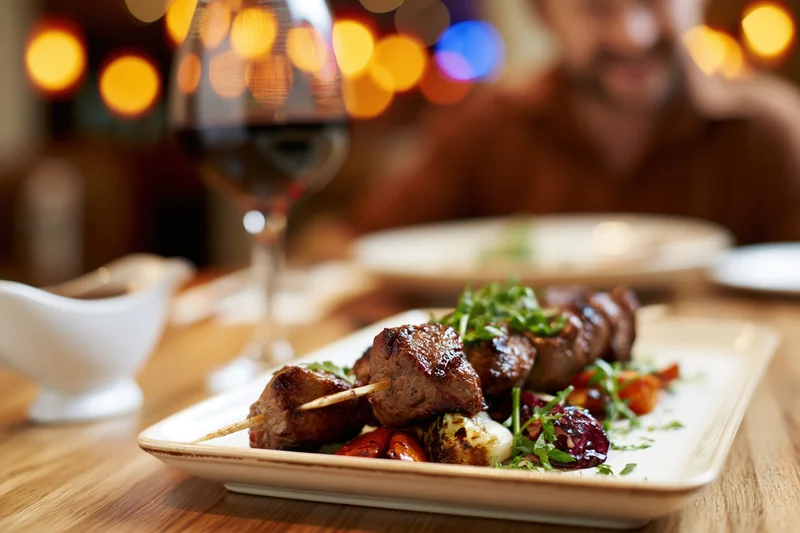 Grilled meat skewers on a white ceramic plate with vegetables and herbs, served at a restaurant table with wine and warm ambient lighting