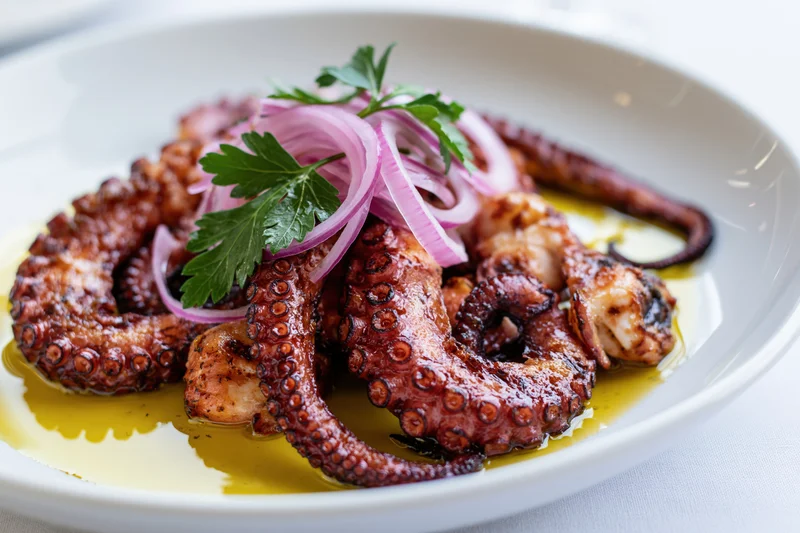 Grilled octopus tentacles arranged on a white ceramic plate with olive oil, red onion, and parsley garnish
