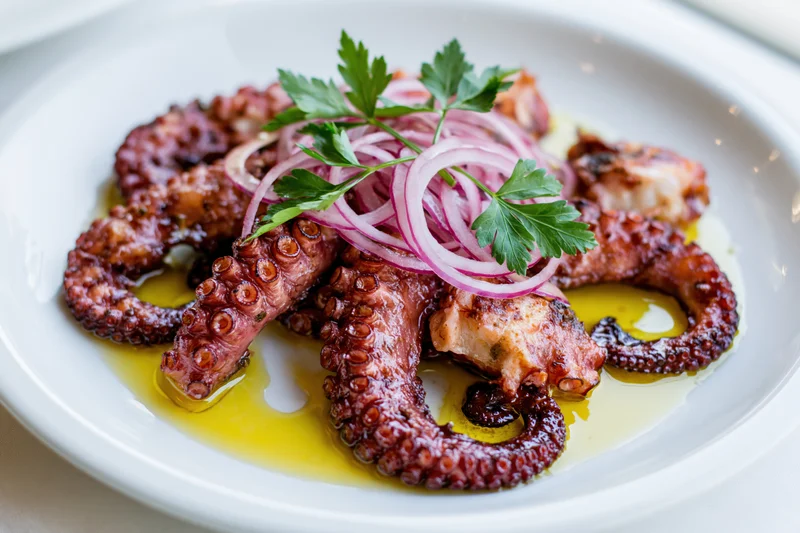 Grilled octopus tentacles arranged on a white ceramic plate with olive oil, red onion, and parsley garnish