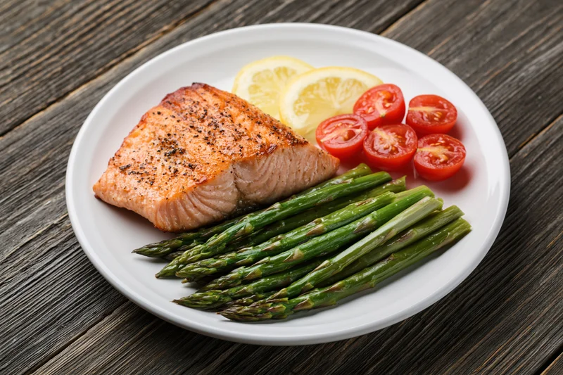 Grilled salmon fillet with steamed asparagus and tomato lemon garnish on white plate over wooden table
