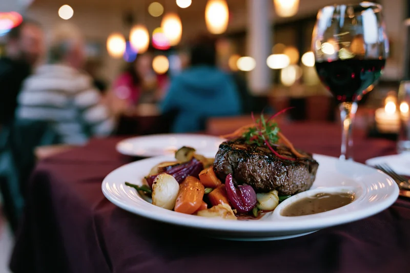 Grilled steak with roasted vegetables and garnish on a white plate at a restaurant table with wine glass and candlelit ambiance