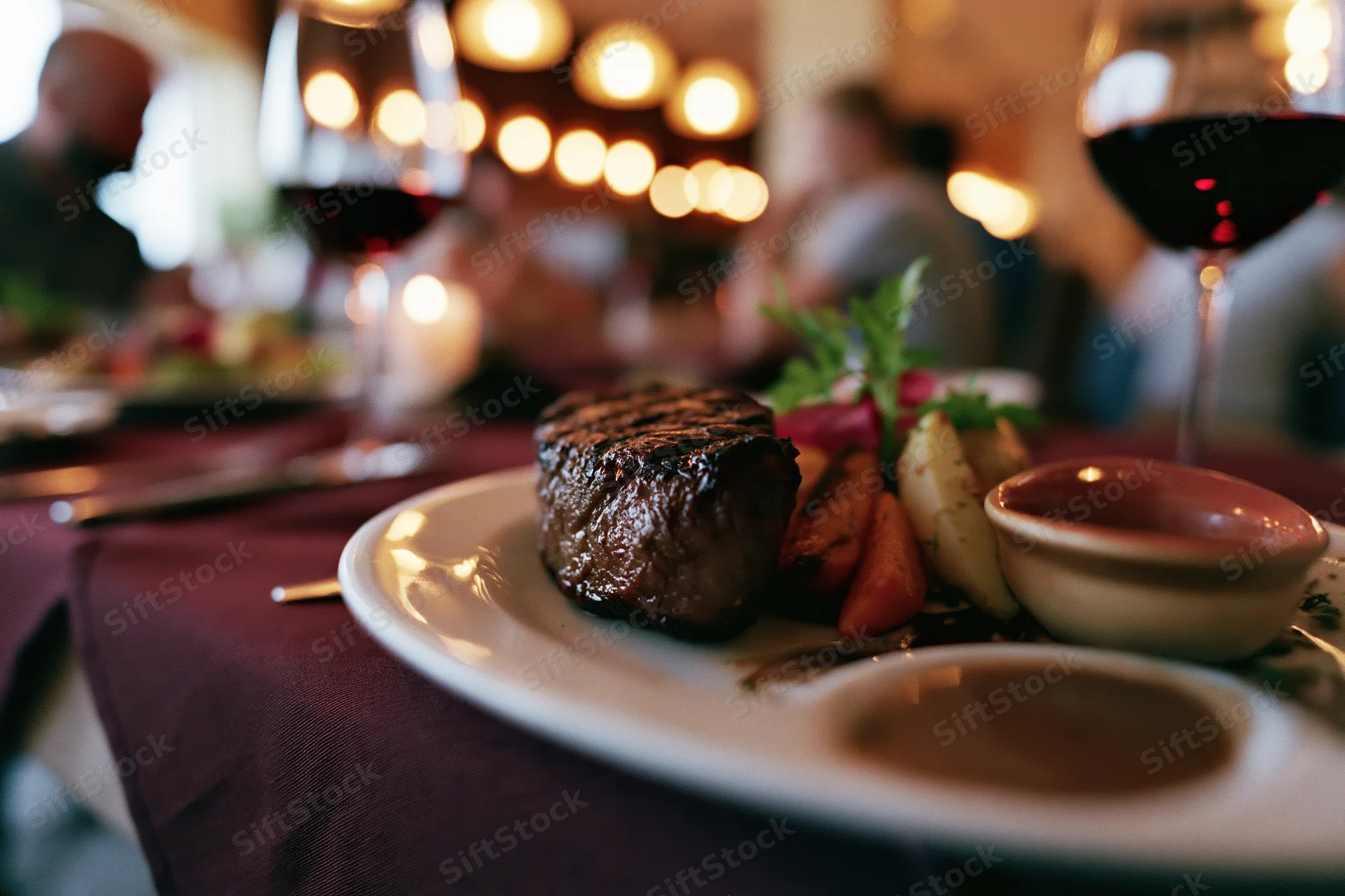 Grilled steak with roasted vegetables and garnish on a white plate at a restaurant table with wine glasses and warm lighting