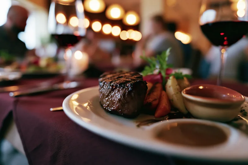 Grilled steak with roasted vegetables and garnish on a white plate at a restaurant table with wine glasses and warm lighting
