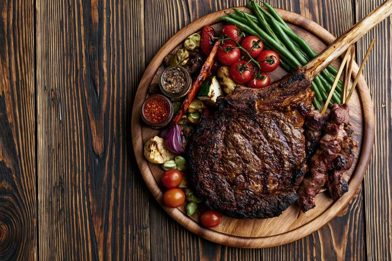 Grilled tomahawk ribeye steak with vegetables and condiments on a round wooden platter