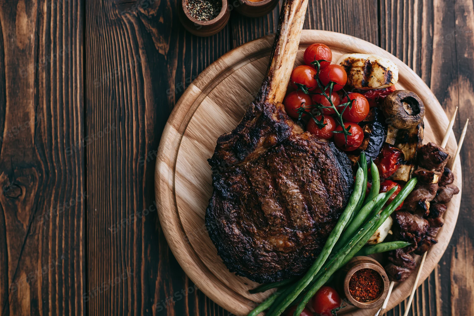 Grilled tomahawk ribeye steak with vegetables and spices on a wooden platter on dark wood table