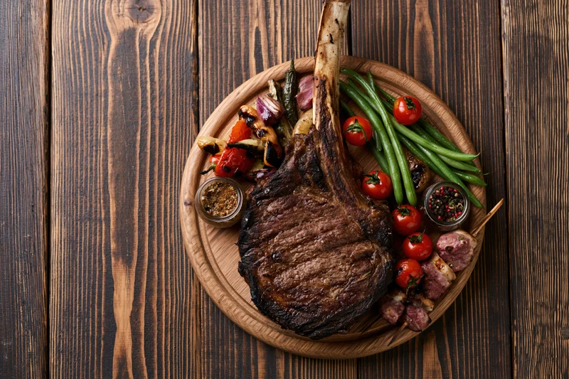 Grilled tomahawk ribeye steak with vegetables and spices on a wooden board over dark wood table