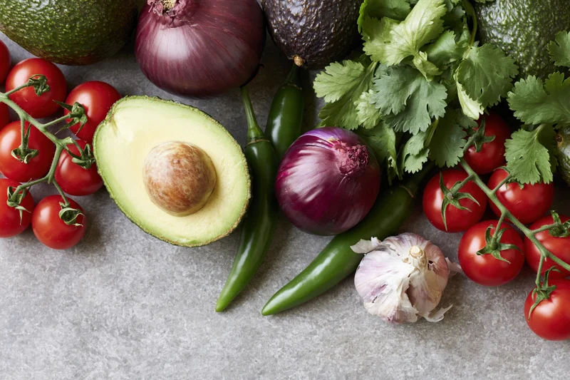 Halved avocado and fresh vegetables arranged on a concrete surface for guacamole preparation