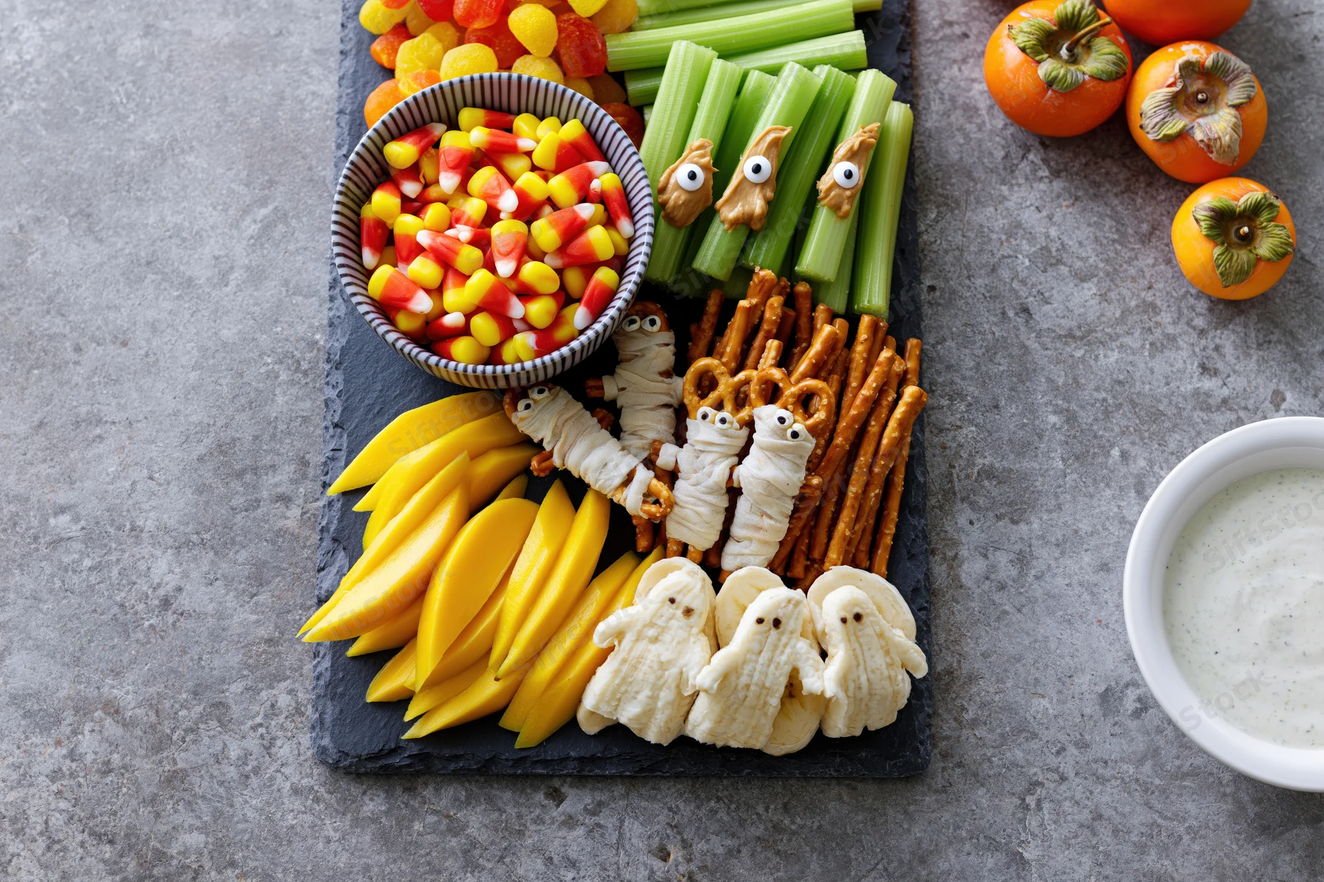 Halloween-themed snack board with candy corn, celery ghosts, pretzel mummies, cheese ghosts, mango slices, persimmons, and dip on slate platter viewed from above