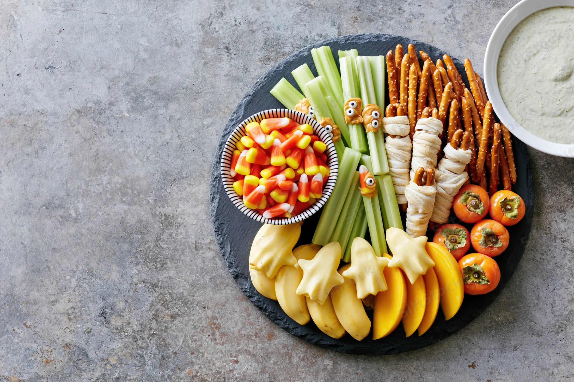 Halloween-themed snack board with celery, pretzels, cheese, fruit and dip arranged on slate platter