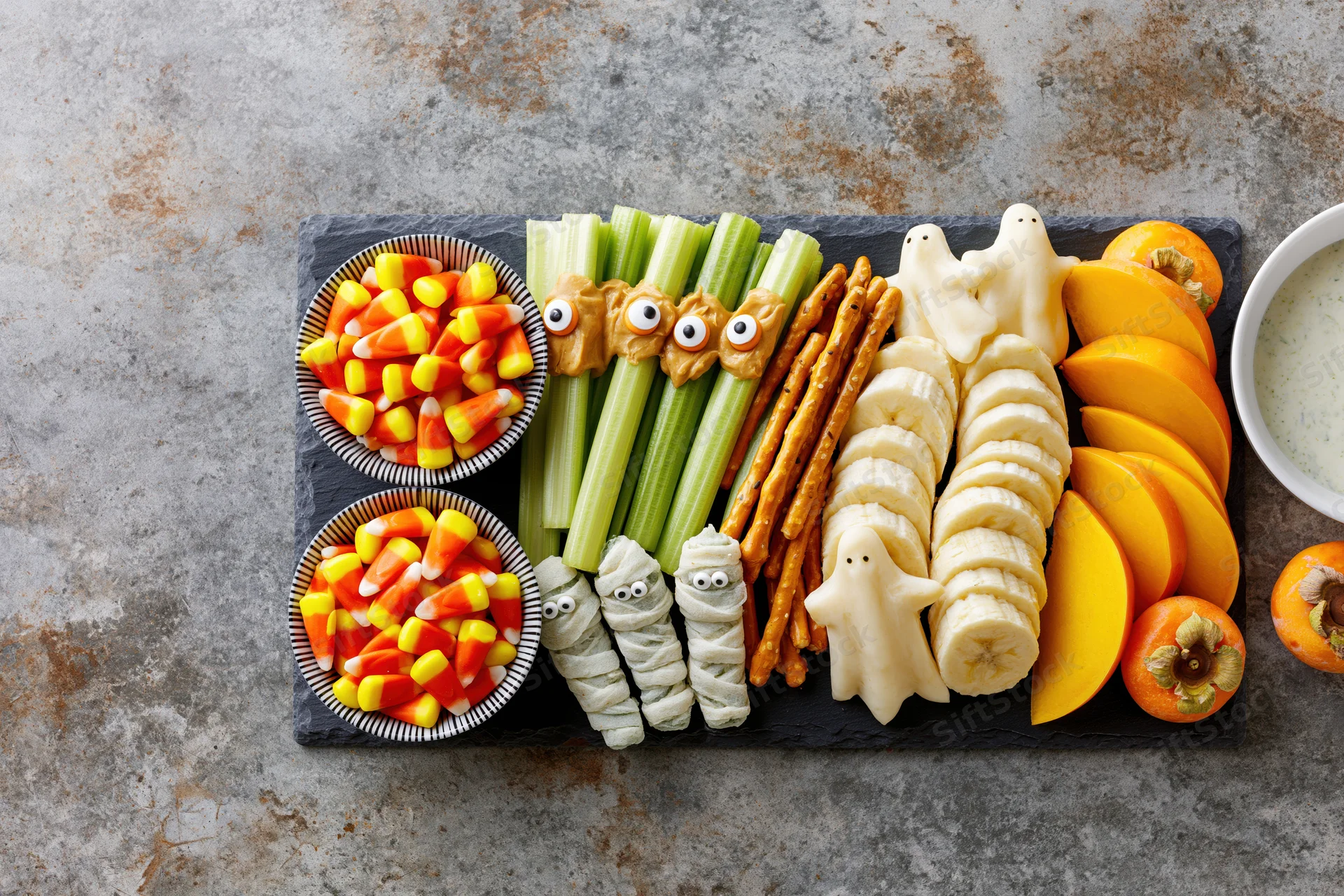 Halloween-themed snack board with celery ghosts, banana mummies, candy corn, and fruit arranged on slate platter