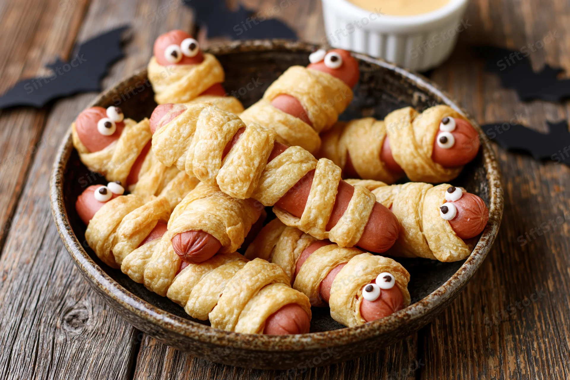 Mummy-themed hot dogs wrapped in baked puff pastry served in a bowl on a wooden table with bat decorations for halloween