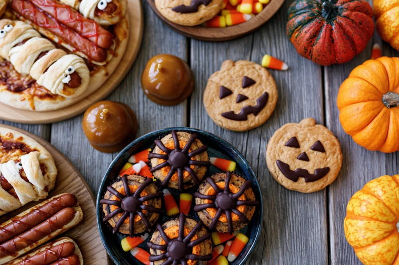 Halloween party food spread with themed cookies, mummy hotdogs, and pumpkins on a rustic wooden table