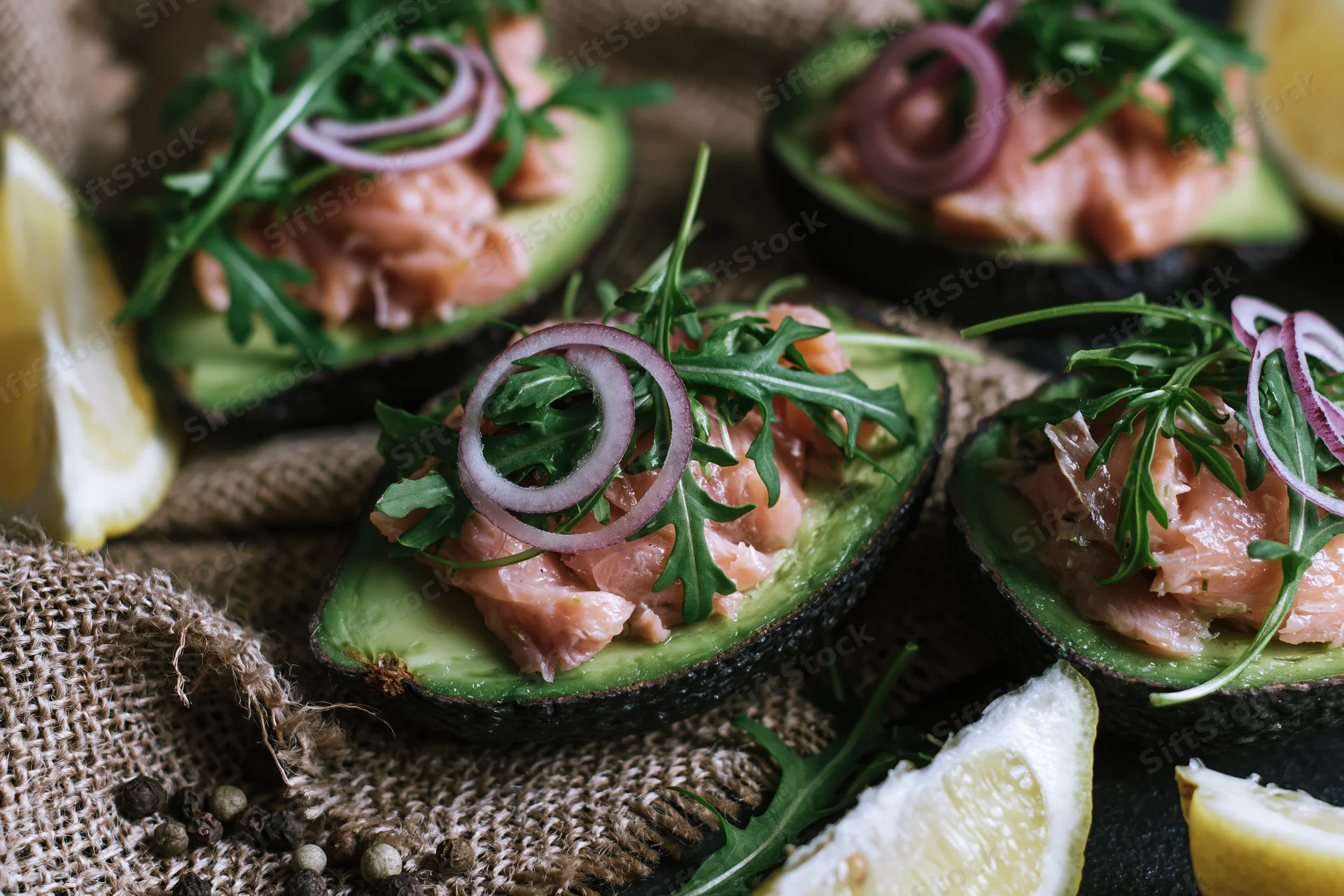 Halved avocados filled with flaked salmon and arugula, topped with red onion rings and lemon wedges on burlap