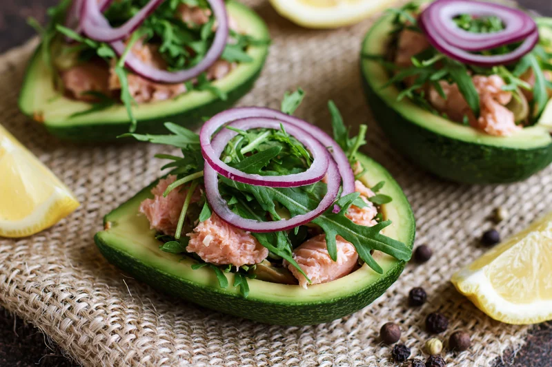 Halved avocados filled with flaked salmon and arugula topped with red onion rings on burlap
