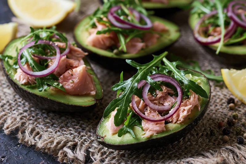 Halved avocados filled with flaked salmon and arugula topped with red onion rings on burlap