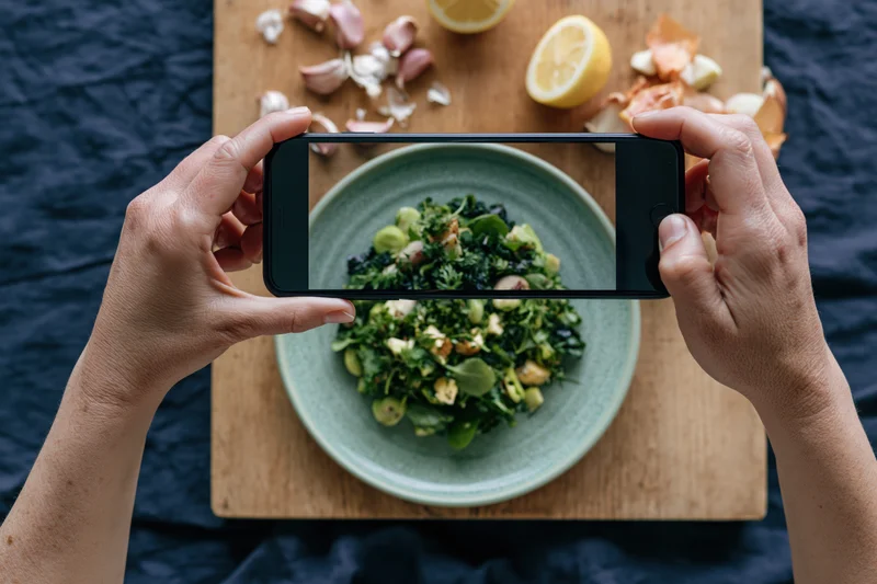Hands holding smartphone horizontally photographing salad on wooden cutting board