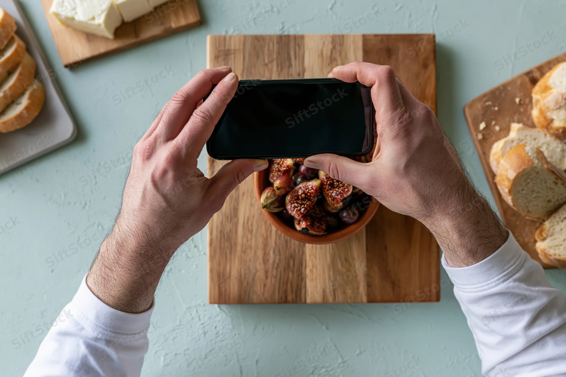 Hands holding smartphone overhead photographing bowl of figs on wooden cutting board with cheese and bread nearby