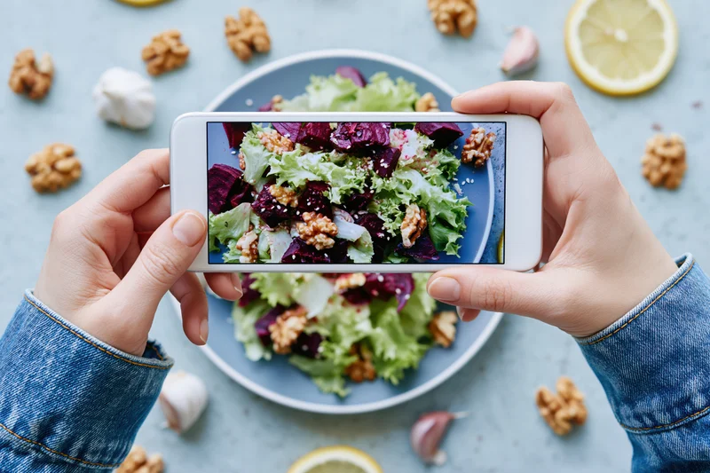 Hands holding smartphone photographing salad on plate with walnuts and lemon slices