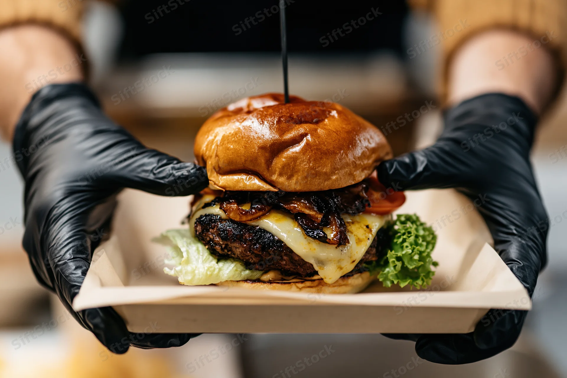 Hands in black nitrile gloves holding a paper tray with a cheeseburger topped with sauce and vegetables