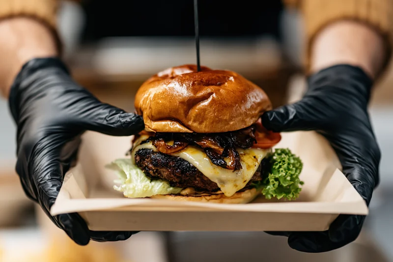 Hands in black nitrile gloves holding a paper tray with a cheeseburger topped with sauce and vegetables