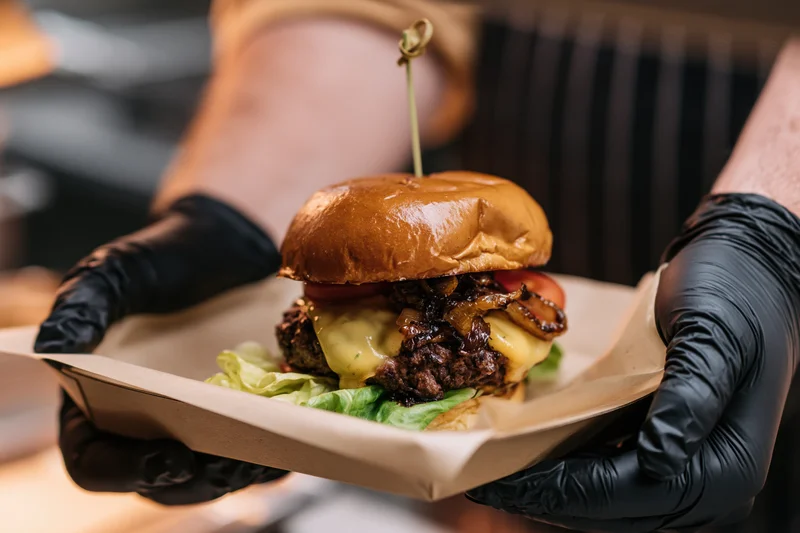 Hands in black nitrile gloves holding a paper tray with a cheeseburger topped with bacon, lettuce, tomato, and cheese, skewered with a toothpick