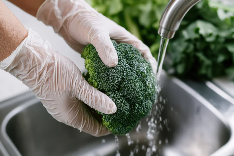 Hands in white gloves rinsing broccoli under running water in a kitchen sink