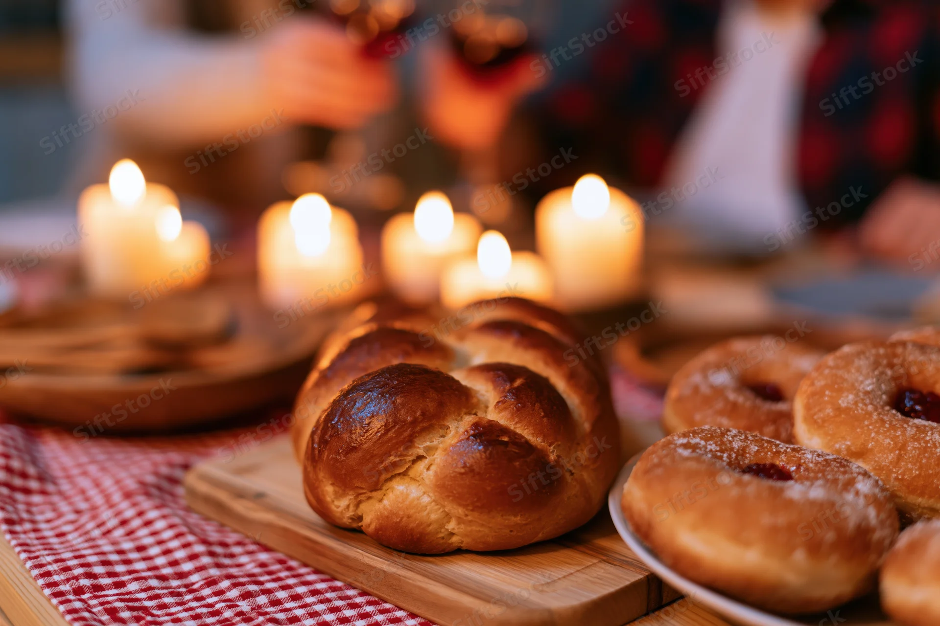 Braided challah on a wooden board with lit candles and sufganiyot on a checkered cloth at a hanukkah dinner table